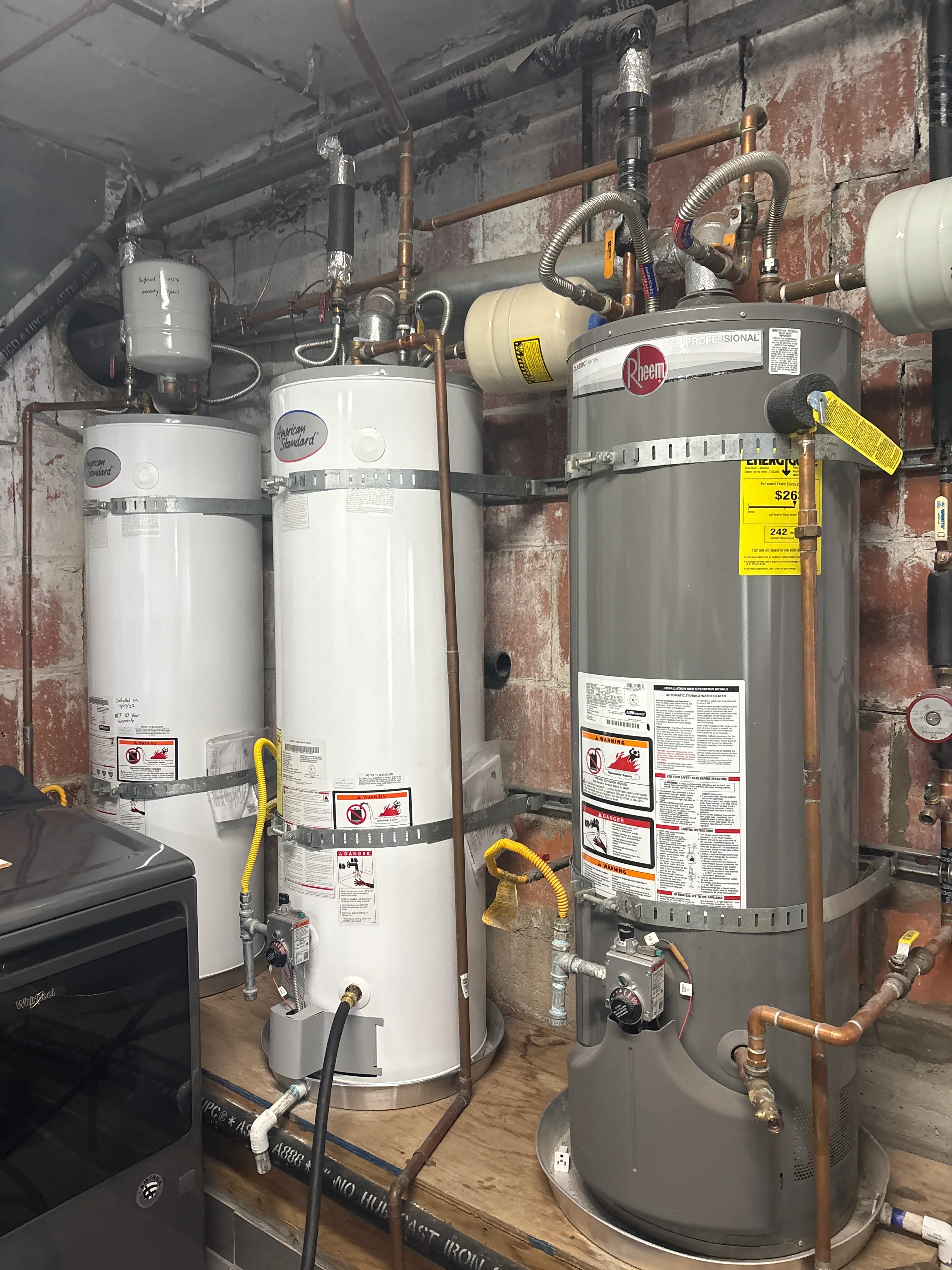 Three water heater tanks with copper piping, flexible connectors, and overhead expansion tank installation